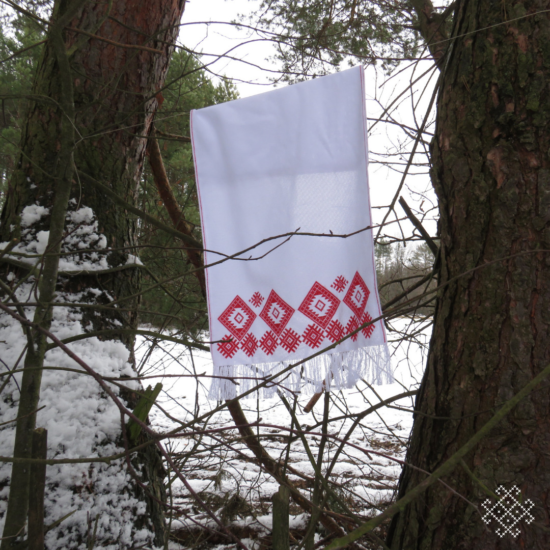 Obraz przedstawia ręcznik dekoracyjny z białego materiału, zawieszony na gałęziach drzew w zimowym, zaśnieżonym lesie. Ręcznik ma tradycyjny haft w czerwonym kolorze, który tworzy geometryczny wzór składający się z rombów oraz motywów ludowych. Dolna krawędź ręcznika jest wykończona białymi frędzlami. Tło sceny jest zimowe, z widocznym śniegiem pokrywającym ziemię oraz gałęzie drzew, co tworzy kontrast z bielą ręcznika i czerwienią haftu.